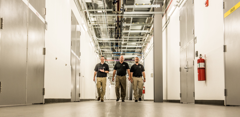 three men walking down an aisle in a large data center warehouse